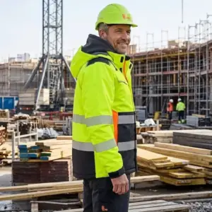 Zimowa kurtka robocza CXS Benson w kolorze żółtym - ujęcie z półprofilu na uśmiechniętym pracowniku w kasku, widoczne poziome pasy odblaskowe, czarne wstawki na dole i szary kaptur - na tle aktywnego placu budowy z rusztowaniami i deskami.