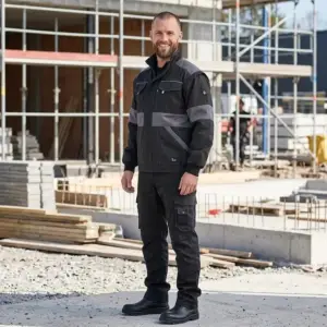 Czarno-szara bluza robocza CXS LUXY Eda - uśmiechnięty mężczyzna w ujęciu całej sylwetki, ubrany w czarną kurtkę z szarymi wstawkami na klatce piersiowej i ramionach oraz czarne spodnie bojówki - na tle słonecznego placu budowy z betonowymi fundamentami i rusztowaniami.