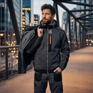 Szaro-czarna bluza dzianinowa Indianapolis na modelu trzymającym kurtkę na ramieniu, widoczny pomarańczowy zamek na klatce piersiowej i czarne wstawki, ujęcie od przodu na tle miejskiej kładki.