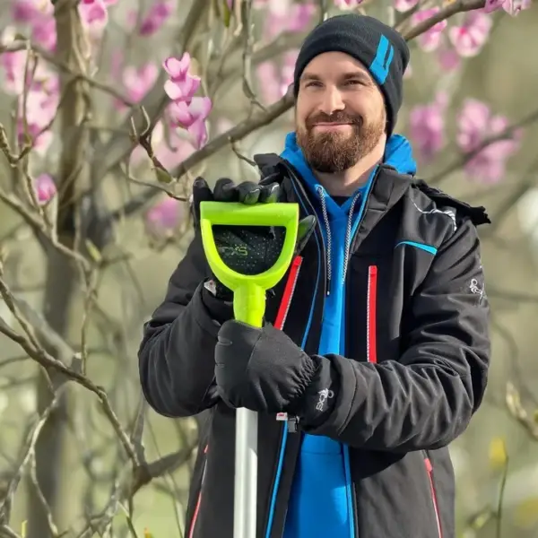 Kurtka Norfolk softshell - uśmiechnięty mężczyzna z brodą ubrany w czarną kurtkę z niebieskimi i czerwonymi detalami, w czapce i rękawicach roboczych, trzymający jaskrawozielony uchwyt łopaty - plenerowe zdjęcie na tle kwitnącej na różowo magnolii.