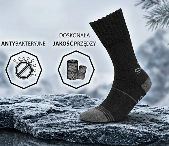 Skarpety TREKKING SILVERWOOL DEODORANT - czarna skarpeta z szarymi wstawkami na pięcie i palcach, ułożona pionowo z profilu. Z lewej strony dwie ikony informacyjne z napisami ANTYBAKTERYJNE i DOSKONAŁA JAKOŚĆ PRZĘDZY - ekspozycja na ośnieżonej skale na tle zimowego krajobrazu.