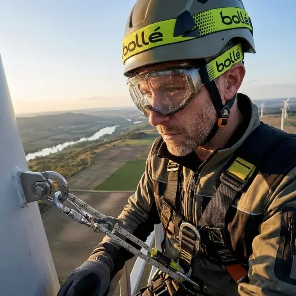 Przezroczysty model BOLLE GLOBE na twarzy pracownika w kasku i uprzęży asekuracyjnej. Zbliżenie z półprofilu na mężczyznę przypiętego liną do białego masztu, w tle rozległy krajobraz z rzeką i turbinami wiatrowymi w świetle zachodzącego słońca.