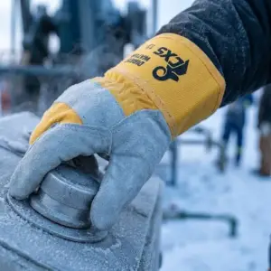 Ocieplane rękawice DINGO WINTER - zbliżenie na dłoń w szaro-żółtej rękawicy z dwoiny bydlęcej, która chwyta oszronione metalowe pokrętło na panelu sterowania - w tle zimowy, zaśnieżony teren przemysłowy z rozmytymi sylwetkami pracowników