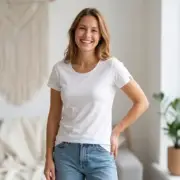 Biała koszulka t-shirt damski Emily z krótkim rękawem i okrągłym dekoltem. Uśmiechnięta modelka w niebieskich dżinsach, widok z przodu w jasnym wnętrzu z makramą i rośliną doniczkową w tle.
