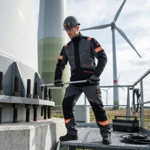 Spodnie ochronne CXS WorkTech-Multi - pracownik w szaro-czarnym uniformie z pomarańczowymi elementami odblaskowymi, stojący na metalowej platformie i dokręcający śrubę długim kluczem - w tle podstawa turbiny wiatrowej i wiatraki na polu